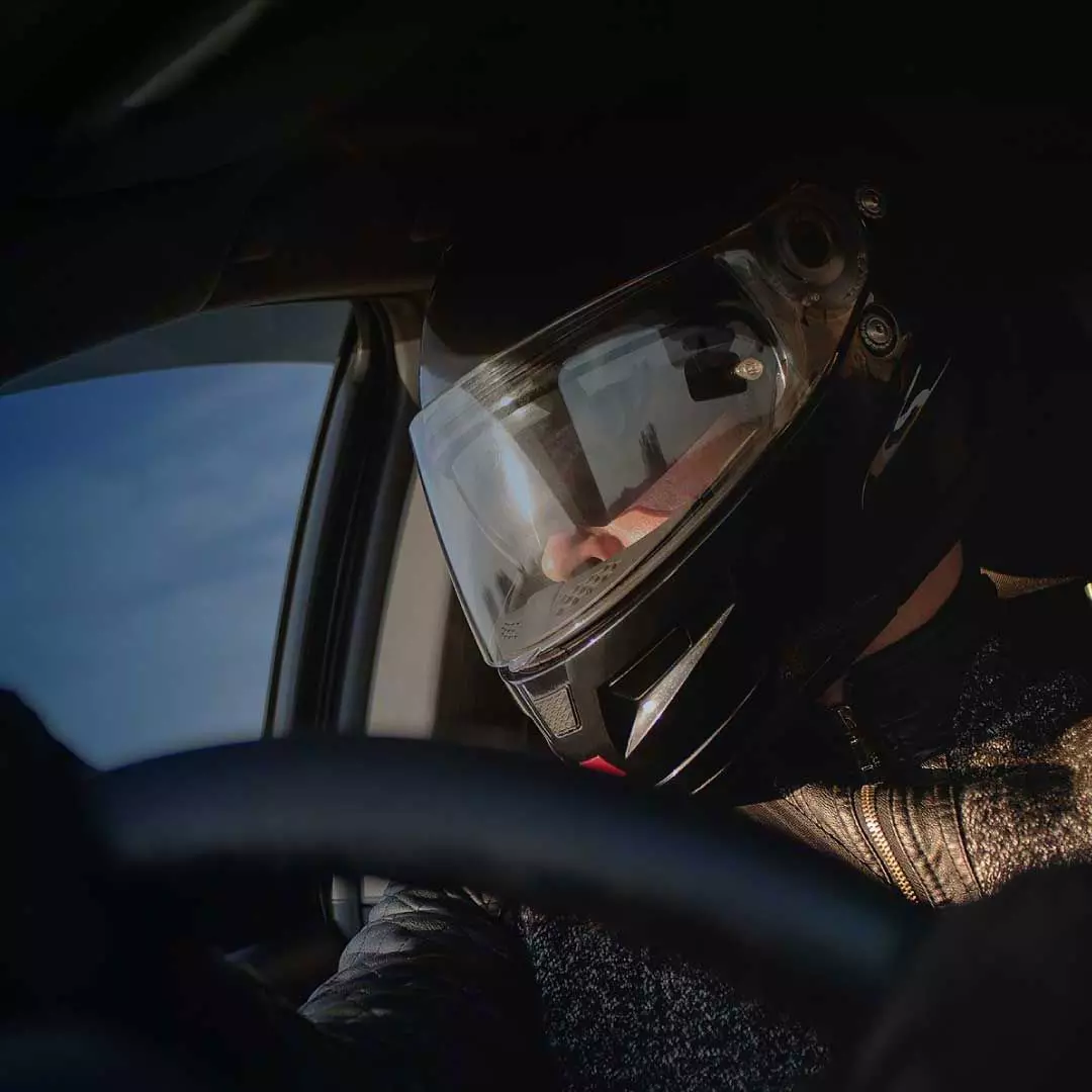 A race car driver grips the wheel, clad in a black helmet with a gleaming face shield.